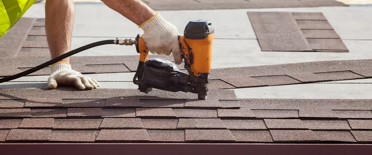 Construction worker putting the asphalt roofing (shingles) with nail gun on a new frame house