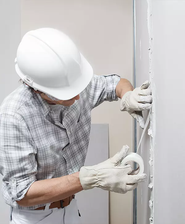 drywall installation, man drywall worker or plasterer putting mesh tape for plasterboard on a wall using a spatula and plaster. Wearing white hardhat, work gloves and safety glasses