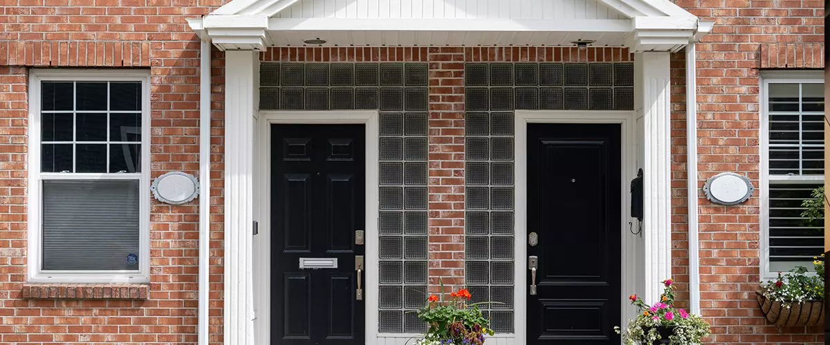 Two black metal doors with flower pots on the steps of duplex houses. It has a red brick wall with white wooden trim. There are double hung windows on both sides of the building entrance